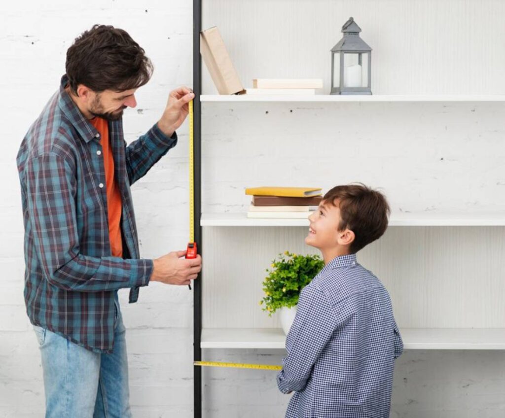A man measures a shelf with a tape measure while a boy stands nearby.