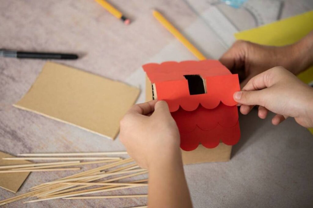 Two hands assemble a colorful paper roof onto a small cardboard house.