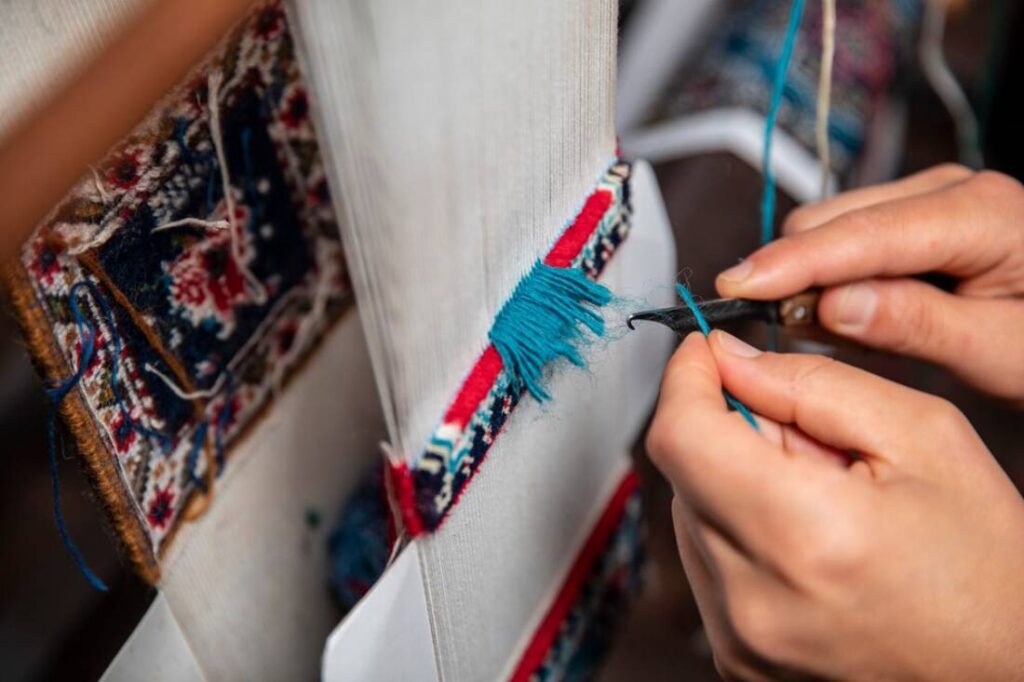 A craftsman's hands work delicately with turquoise threads on a weaving loom