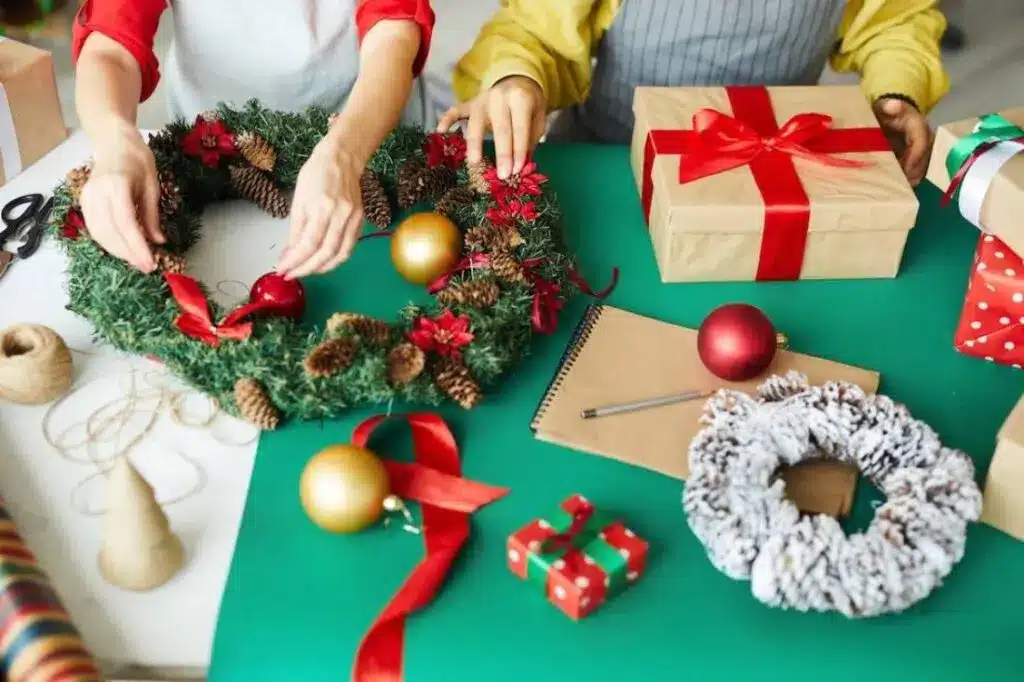 Two hands work on a festive Christmas wreath amidst wrapped gifts.