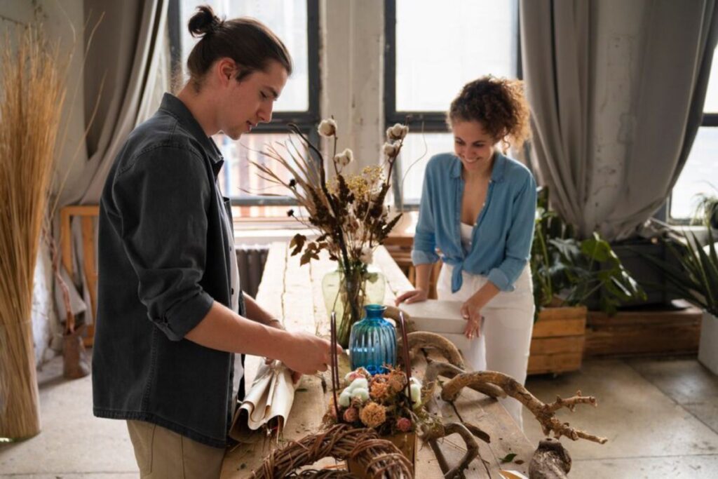 Two people are arranging dried flowers and branches on a wooden table in a bright room