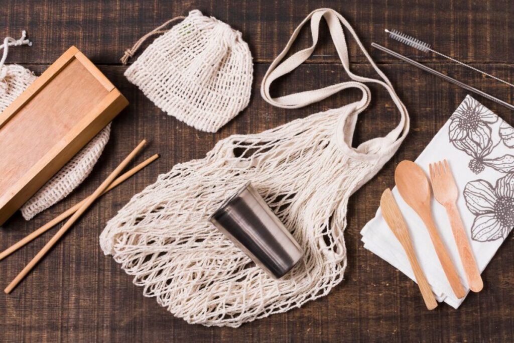 An assortment of eco-friendly items: a woven bag, stainless cup, wooden utensils, and cloth napkin arranged on a wooden surface.