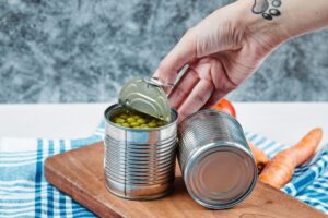 A person opening a can of green peas on a wooden cutting board, with carrots in the background.